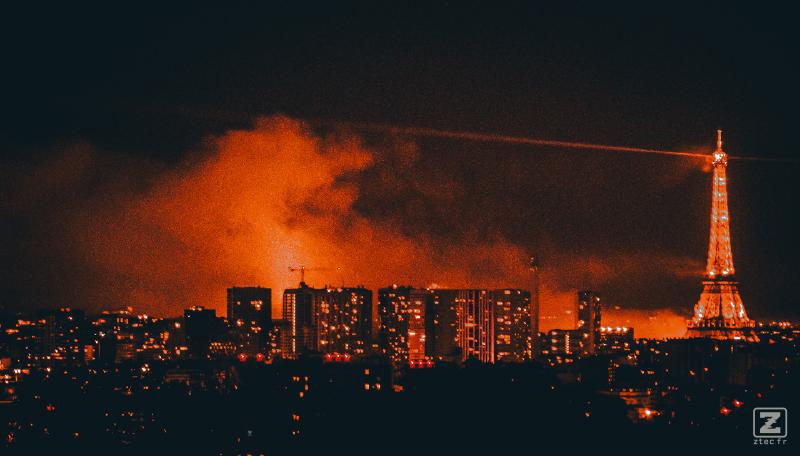 Paris from afar with the Eiffel tower. Clouds of smoke lighted in orange is visible behind buildings and the tower.