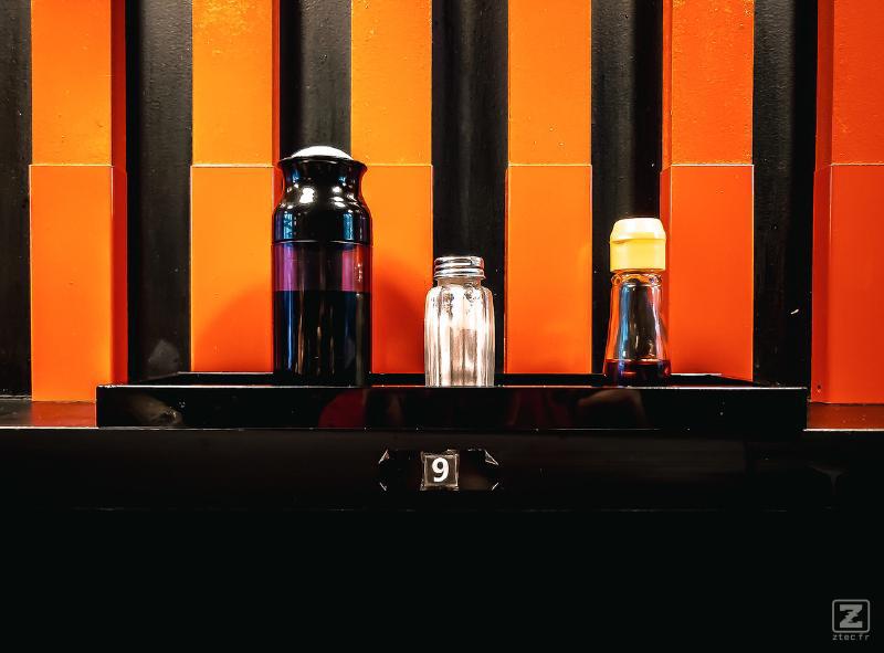 Sauces soja et condiment sur le comptoir avec le mur juste derrière composé de bandes rouge/orange et noir. Le numéro 9 est visible sur la tranche du comptoir
https://www.flickr.com/photos/ztec/53160273843/