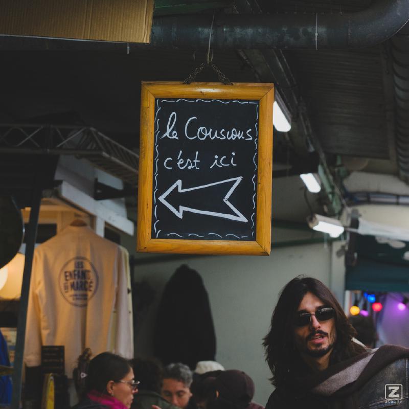 A sign with an arrow pointing left with the mention "Le couscous c'est ici". A guy with long hair and sunglasses contrast the scene on the right. 