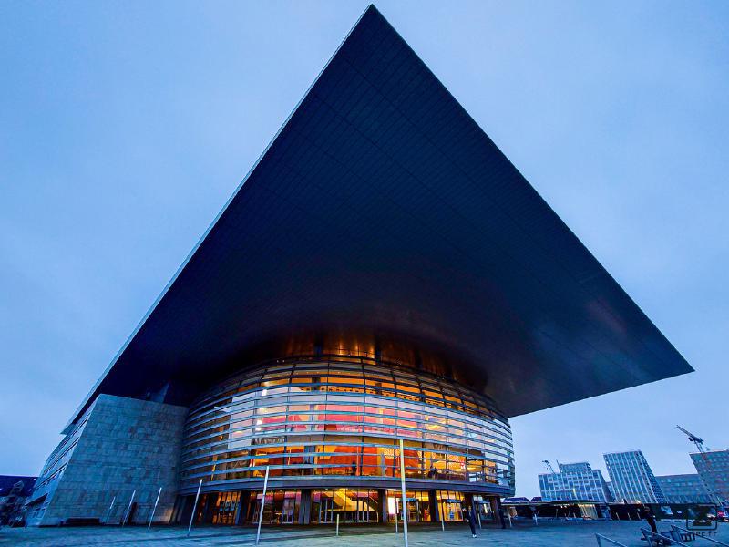 Picture of the Copenhagen opera. A bubble with glowing orange from inside (actually wood) right below a flat square that act as a roof, but go way longer outside the bubble. The picture is deforming the square making one corner pointing to the top border of the picture. Main colors are blue shared everywhere, except the bubble interior.  
https://www.flickr.com/photos/ztec/53132985520/
