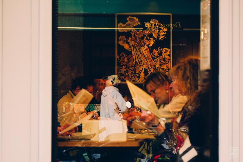 A peek inside a restaurant though a window, with a lady in the glass reflection.
