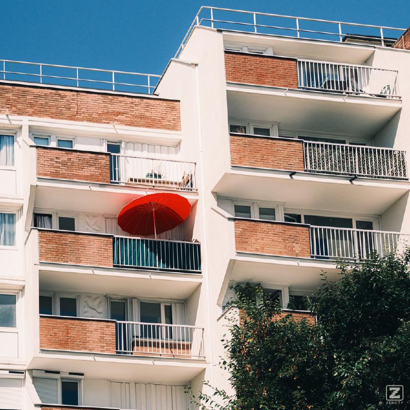 From the balconies of a building, a red parasol juts out from one of them.