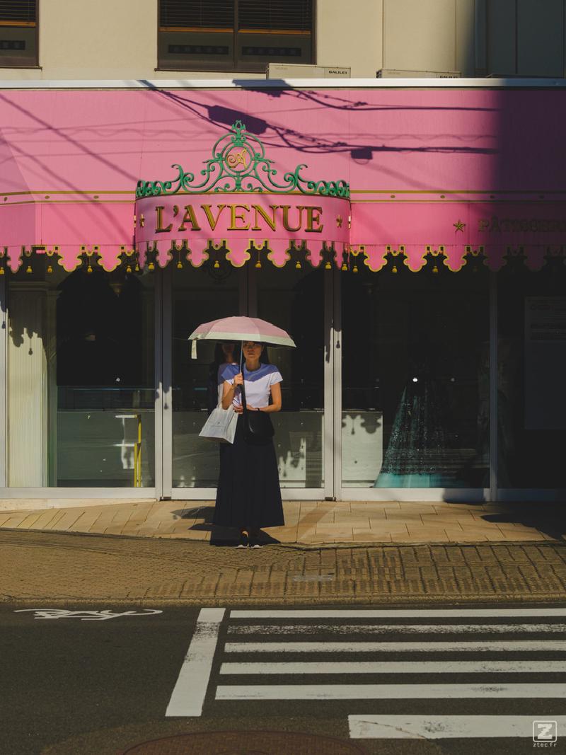A lady is waiting at a crossing with a white and pink umbrella protecting her from the sun