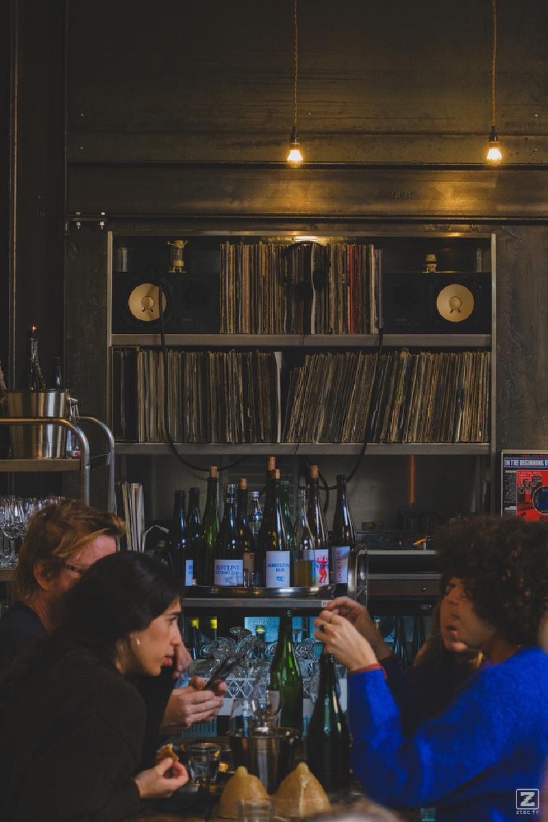 A restaurant where people are talking at the table. In the background there is Vinyles shelfs with speakers. 