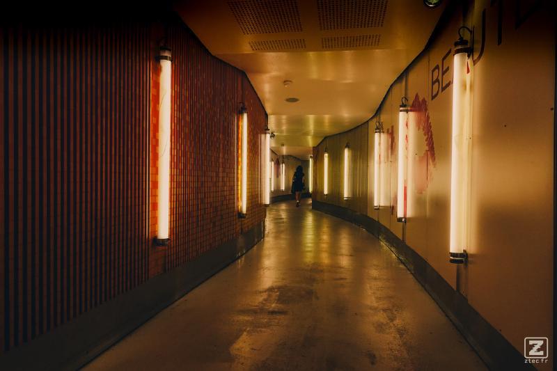 A corridor with neon lights on each walls (vertical). The corridor is zigzaging. We can see a woman walking in the end.