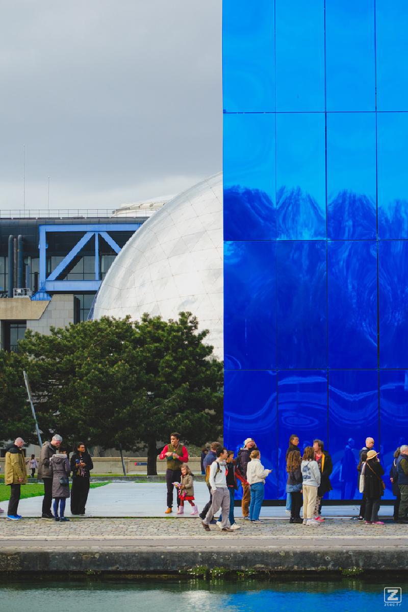 A blue building in front of a mirror sphere, with people waiting in line in front.