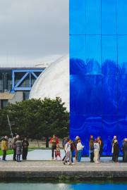 A blue building in front of a mirror sphere, with people waiting in line in front.