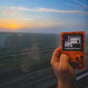 Picture of an orange Gameboy pocket with the window of an highspeed train behind with a flat lanscape. 