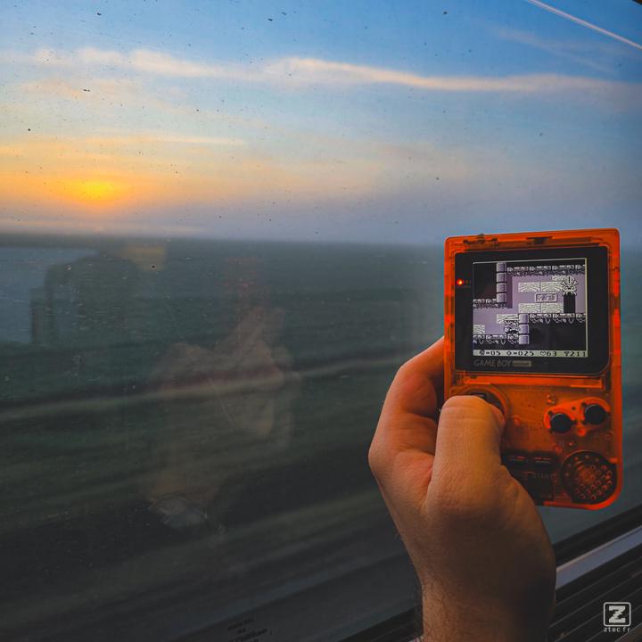 Picture of an orange Gameboy pocket with the window of an highspeed train behind with a flat lanscape. 