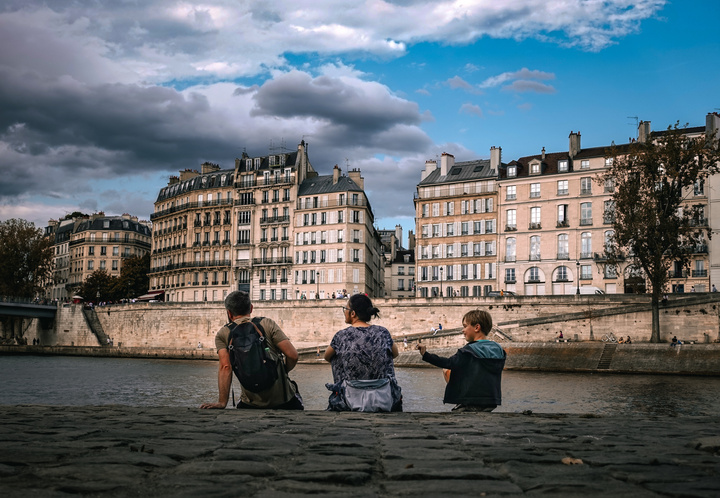 3 person sited on the river border. The river is the Seine in Paris. We can see the typical Parisian building on the other side of the river. 
There is a man, a woman and a child. The child is fidgeting with a feather. 
https://www.flickr.com/photos/ztec/53160266258/