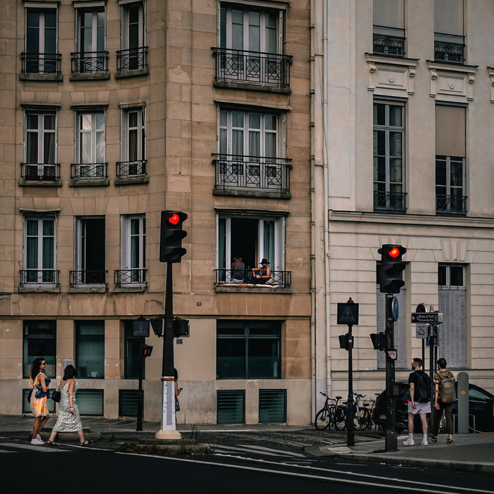 A Parisian street with two red stop light. First floor, a guy is sitting on the (small) balcony with a black hat looking in the direction of the camera taking the picture. Two girls walk by on the left, and two boy walk by on the right
https://www.flickr.com/photos/ztec/53159776726/