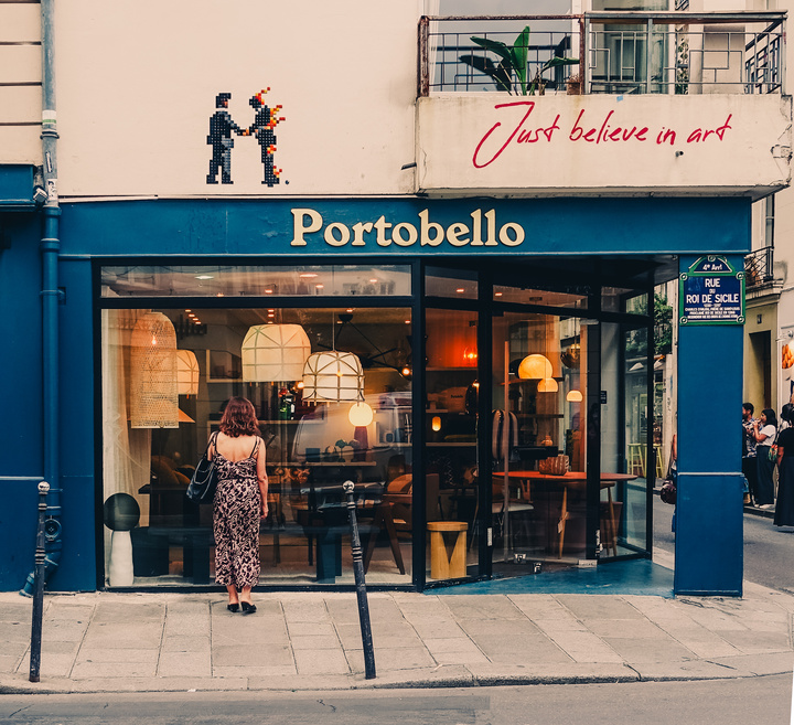 A girl is looking into the window of a store. There is design furniture in the store. The store is names &quot;Portobello&quot;
Right above the store, a Mosaic display two man shaking hand, one is in fire (pink Floyd cover?). Right to it, in a different style, is written &quot;Just believe in art&quot;. 
https://www.flickr.com/photos/ztec/53159982904/