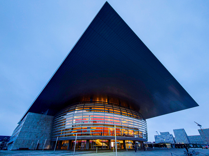 Picture of the Copenhagen opera. A bubble with glowing orange from inside (actually wood) right below a flat square that act as a roof, but go way longer outside the bubble. The picture is deforming the square making one corner pointing to the top border of the picture. Main colors are blue shared everywhere, except the bubble interior.  
https://www.flickr.com/photos/ztec/53132985520/