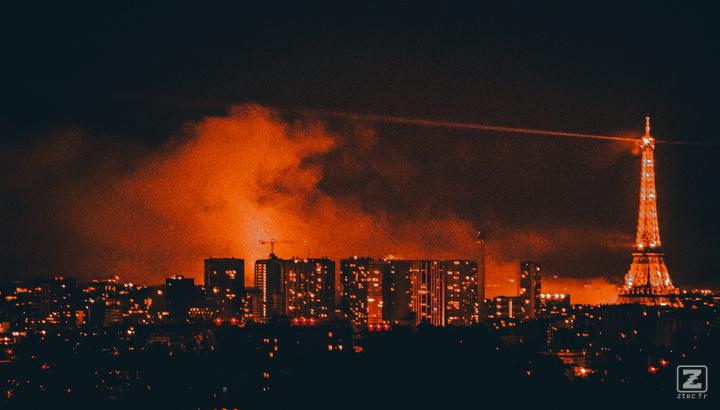 Paris from afar with the Eiffel tower. Clouds of smoke lighted in orange is visible behind buildings and the tower.