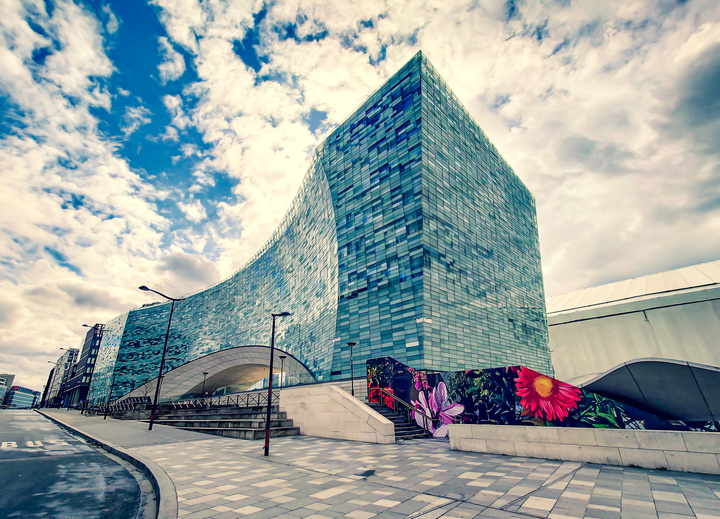 Modern building made of glass (shade of blue). A blue sky with white clouds behind. The building has an arch in the middle, you can walk under it. On the right, one wall is black with colorfull flower painted on it, the streetart that make all the picture shine
https://www.flickr.com/photos/ztec/53132575311/