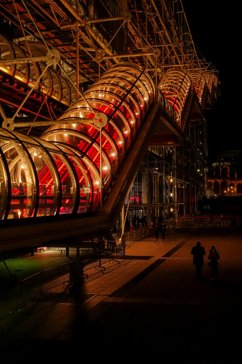 At night, a stair inside a tube that goes up. It look like an airport transit hallway. There is two silhouette below the stairs.

