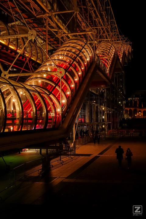 At night, a stair inside a tube that goes up. It look like an airport transit hallway. There is two silhouette below the stairs.