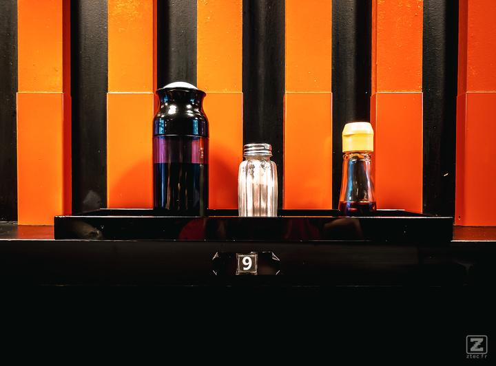 Sauces soja et condiment sur le comptoir avec le mur juste derrière composé de bandes rouge/orange et noir. Le numéro 9 est visible sur la tranche du comptoir
https://www.flickr.com/photos/ztec/53160273843/