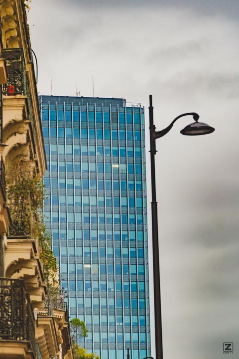 Hausmanian building, glass building, and a light pole in the sky, all aligned.