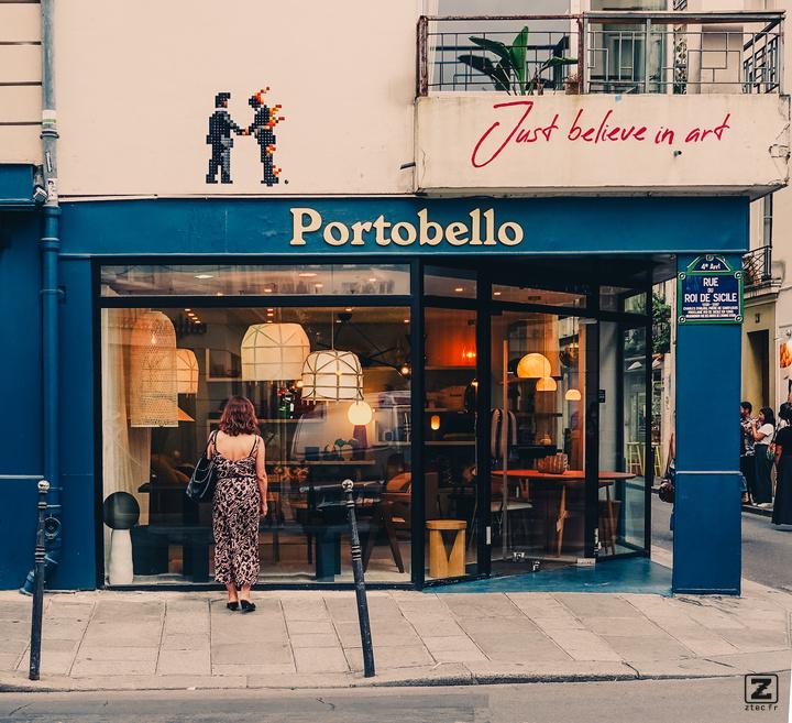 A girl is looking into the window of a store. There is design furniture in the store. The store is names "Portobello"
Right above the store, a Mosaic display two man shaking hand, one is in fire (pink Floyd cover?). Right to it, in a different style, is written "Just believe in art". 
https://www.flickr.com/photos/ztec/53159982904/