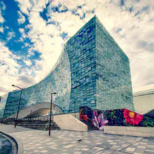 Modern building made of glass (shade of blue). A blue sky with white clouds behind. The building has an arch in the middle, you can walk under it. On the right, one wall is black with colorfull flower painted on it, the streetart that make all the picture shine
https://www.flickr.com/photos/ztec/53132575311/