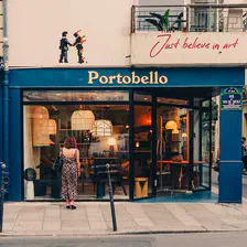 A girl is looking into the window of a store. There is design furniture in the store. The store is names &quot;Portobello&quot;
Right above the store, a Mosaic display two man shaking hand, one is in fire (pink Floyd cover?). Right to it, in a different style, is written &quot;Just believe in art&quot;. 
https://www.flickr.com/photos/ztec/53159982904/