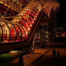 At night, a stair inside a tube that goes up. It look like an airport transit hallway. There is two silhouette below the stairs.
