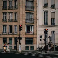 A Parisian street with two red stop light. First floor, a guy is sitting on the (small) balcony with a black hat looking in the direction of the camera taking the picture. Two girls walk by on the left, and two boy walk by on the right
https://www.flickr.com/photos/ztec/53159776726/