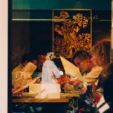 A peek inside a restaurant though a window, with a lady in the glass reflection.