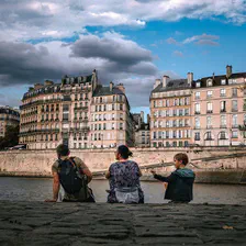 3 person sited on the river border. The river is the Seine in Paris. We can see the typical Parisian building on the other side of the river. 
There is a man, a woman and a child. The child is fidgeting with a feather. 
https://www.flickr.com/photos/ztec/53160266258/