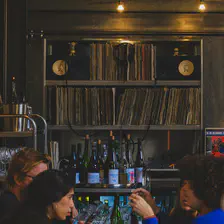 A restaurant where people are talking at the table. In the background there is Vinyles shelfs with speakers. 