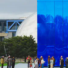 A blue building in front of a mirror sphere, with people waiting in line in front.