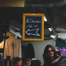 A sign with an arrow pointing left with the mention "Le couscous c'est ici". A guy with long hair and sunglasses contrast the scene on the right. 