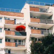 From the balconies of a building, a red parasol juts out from one of them.