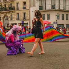 A woman walking from left to right, with a giant LGBT+ flag behind. An another person is squatting on the left, wearing full purple clothes. 