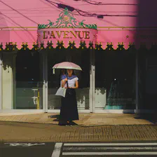 A lady is waiting at a crossing with a white and pink umbrella protecting her from the sun